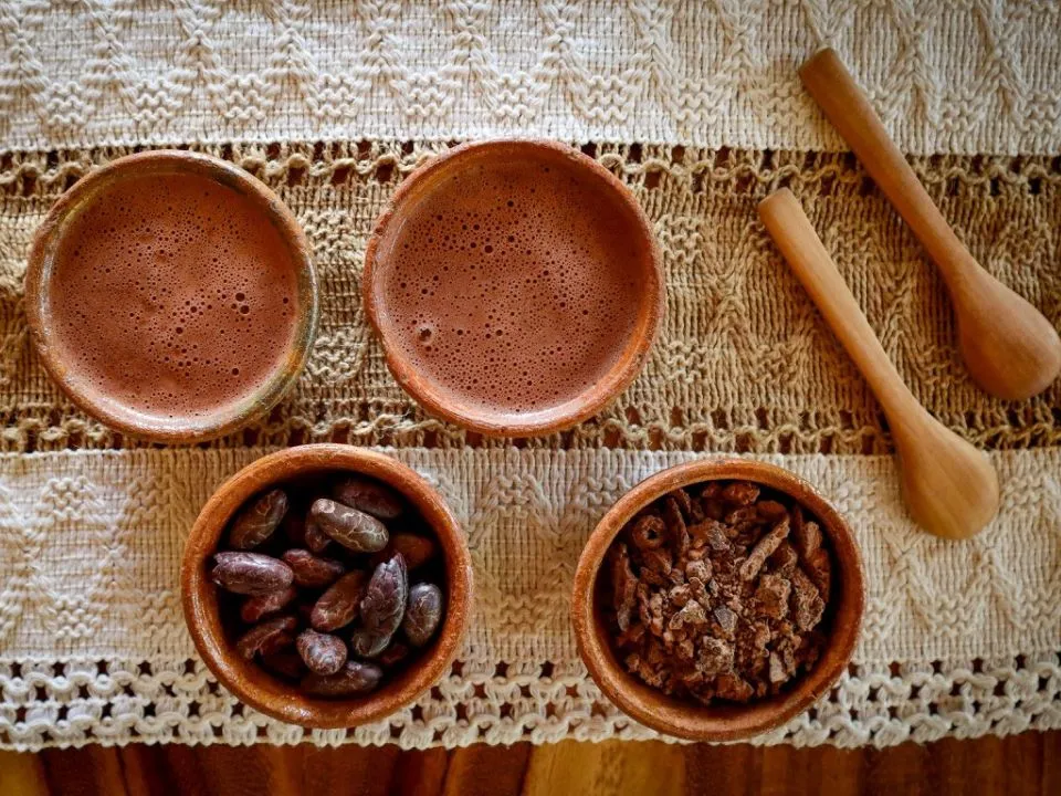 Two cups of frothy ceremonial cacao beside bowls of cacao beans and nibs on a woven cloth